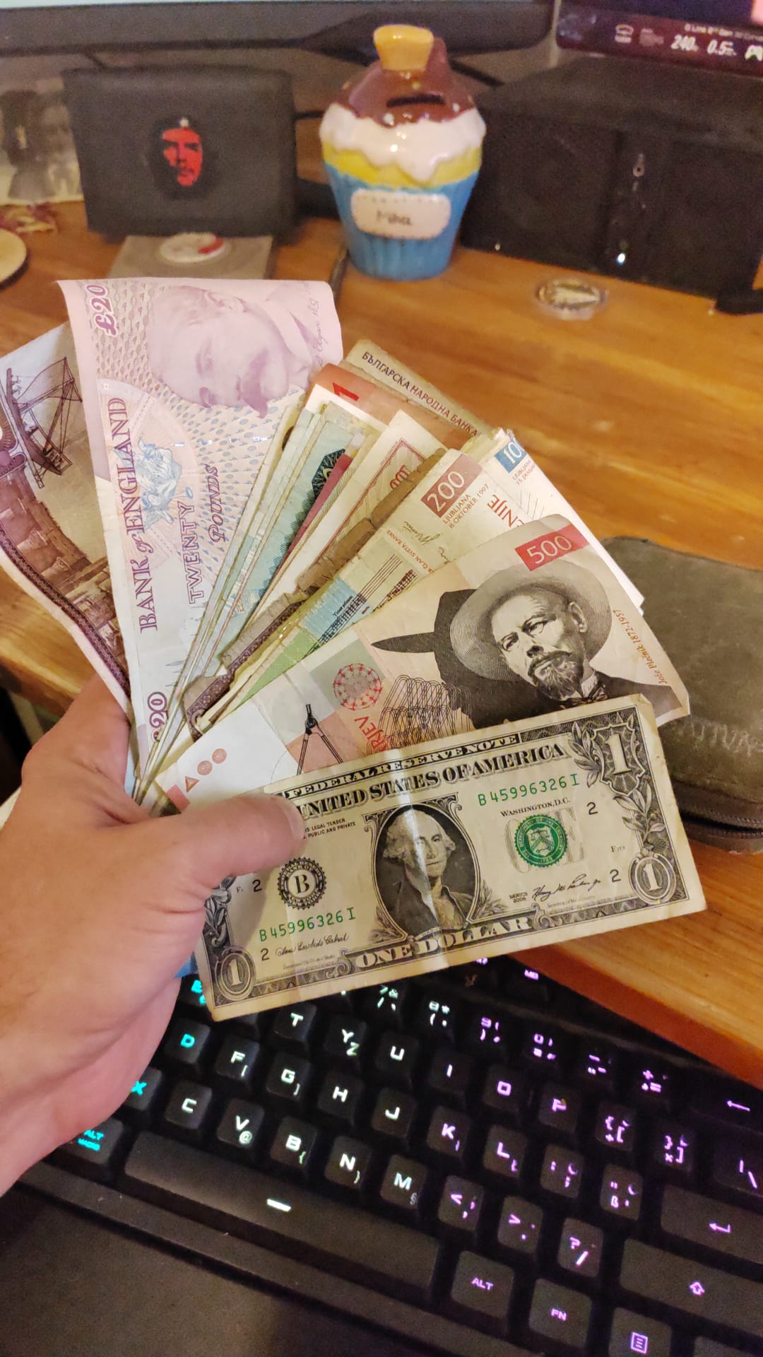 A hand fanning out a US 1 dollar bill, a Slovenian 500 Tolar with Jože Plečnik, a British 20 pound note and other world banknotes — three eras of monetary dominance in one frame.